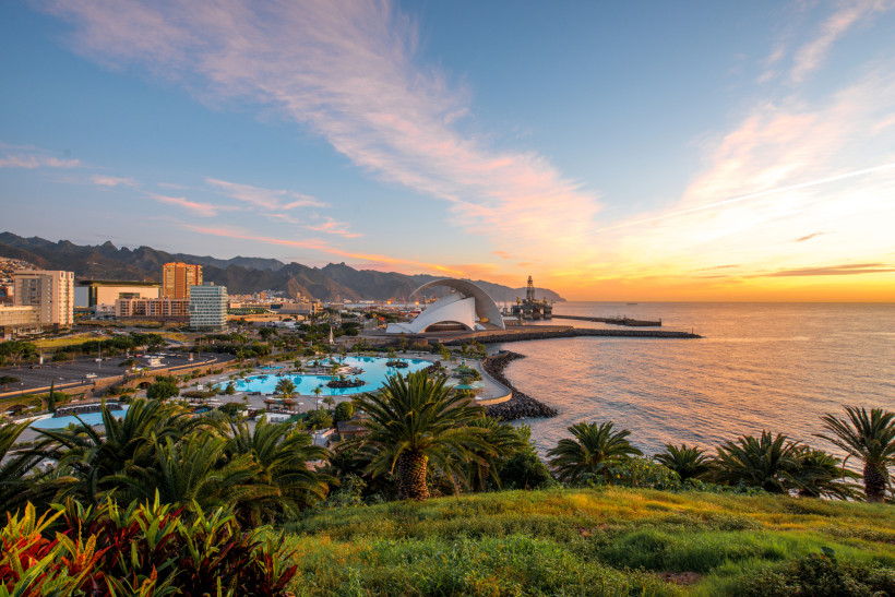 Sonnenuntergang über Santa Cruz de Tenerife mit Blick auf das Auditorium Panorama von Santa Cruz de Tenerife bei Sonnenuntergang. Im Vordergrund tropische Gärten und Pools, dahinter das moderne, muschelförmige Auditorium von Teneriffa am Meer. Sanft beleuchteter Himmel in Orange- und Rosatönen, rechts der Atlantik, im Hintergr
