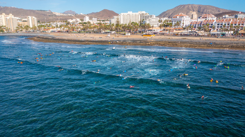 Teneriffa - Playa Honda Surfer im Meer, dahinter breiter Sandstrand, Promenade mit Palmen und Hotels vor den Bergen.
