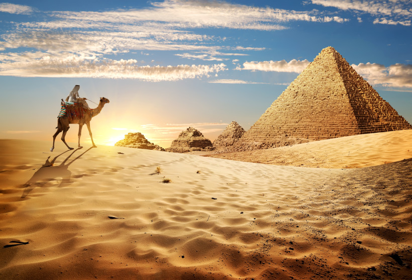 Ägypten Das Bild zeigt eine weite Wüstenlandschaft bei Sonnenuntergang. Im Vordergrund zieht ein Mann in traditioneller arabischer Kleidung auf einem Kamel durch den weichen, goldfarbenen Sand. Die Sonne geht hinter ihm unter und wirft lange Schatten. Im Hintergr