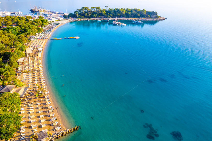 Luftaufnahme einer Badebucht in Kemer mit Strandliegen, türkisblauem Wasser, Booten und Pinien an der Türkischen Riviera