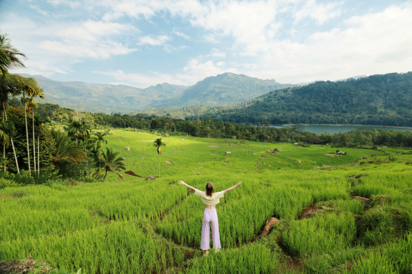 Sri Lanka Frau mit ausgestreckten Armen steht auf einem schmalen Pfad inmitten leuchtend grüner Reisterrassen. Im Hintergrund erstrecken sich dichte Wälder, ein See und ein Gebirge unter blauem Himmel. Die Szene strahlt Ruhe, Naturverbundenheit und tropische Weite