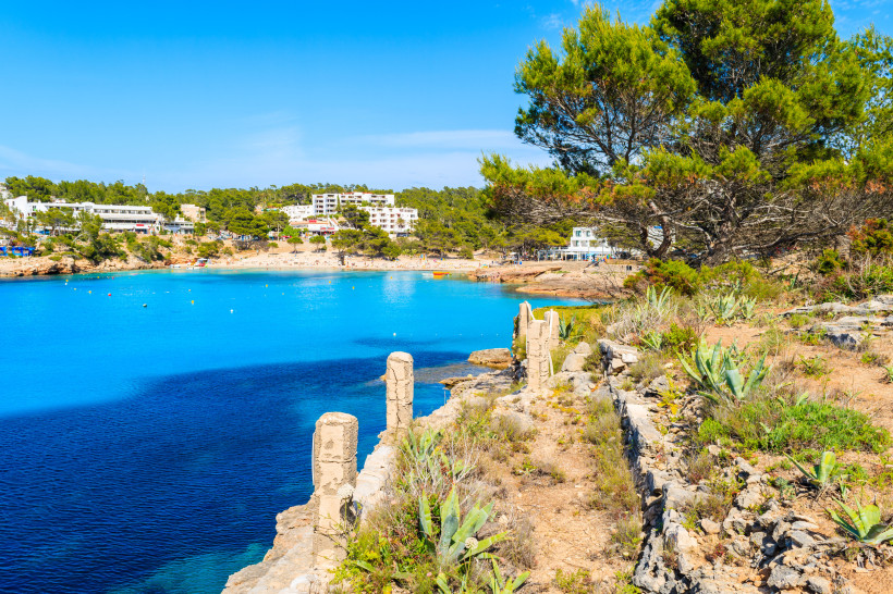 Ibiza - Portinatx Küstenansicht eines Ferienortes im Norden von Ibiza. Im Vordergrund Felsen, Kakteen und Pinien, dahinter türkisblaues Meer und Sandstrand mit Hotels und Ferienanlagen.