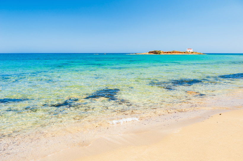 Malia Beach mit flachem, klarem Meerwasser und Sandstrand, im Hintergrund kleine Insel mit weißem Gebäude