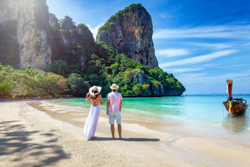 Thailand Das Bild zeigt ein Paar in sommerlicher Kleidung, das entspannt an einem tropischen Sandstrand steht. Die Frau trägt ein weißes, luftiges Kleid und einen Sonnenhut, der Mann helle Shorts, ein rosa Shirt und ebenfalls einen Hut. Beide blicken auf das ruhig