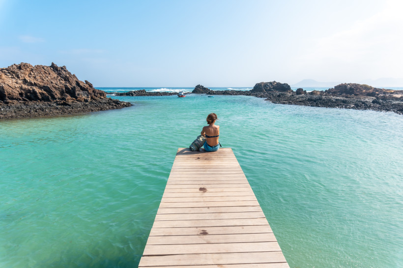 Isla de Lobos Naturpools – Holzsteg am türkisfarbenen Wasser bei Fuerteventura Frau sitzt auf einem Holzsteg an den Naturpools der Isla de Lobos, umgeben von türkisfarbenem Wasser und Lavafelsen vor Fuerteventura.