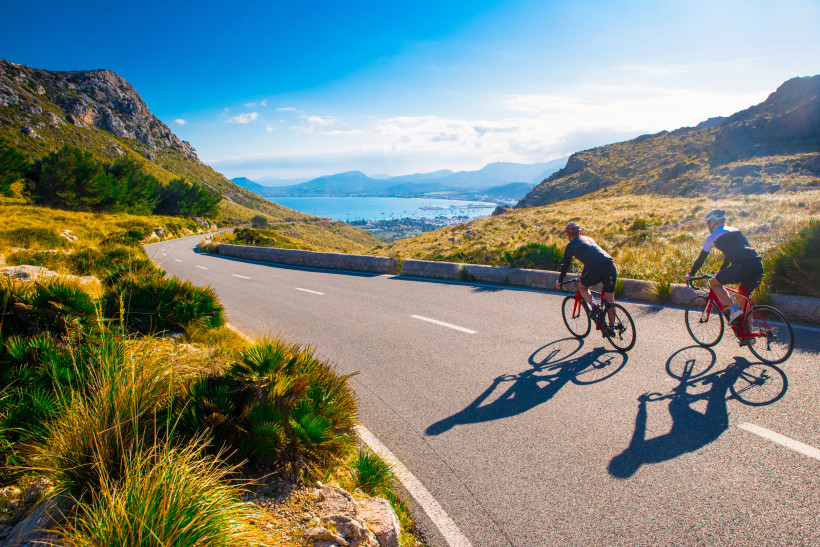 Mallorca Zwei Rennradfahrer fahren auf einer kurvigen, asphaltierten Panoramastraße durch eine bergige Landschaft mit Blick auf eine Bucht. Die Umgebung ist geprägt von goldenen Gräsern, Felsen und einzelnen Büschen. Im Hintergrund erstreckt sich das Meer mit eine