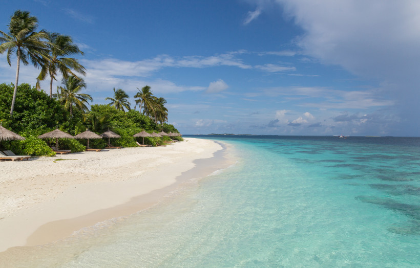 Sandstrand an einer Atollinsel auf den Malediven mit Palmen, Sonnenschirmen und türkisfarbenem Meer