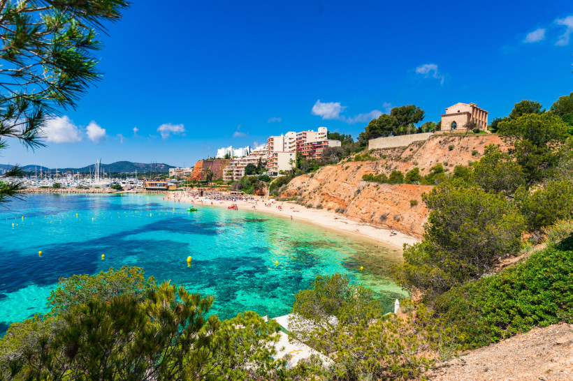 Blick auf die Bucht von Portals Nous mit Platja de l’Oratori, türkisfarbenem Wasser, Sandstrand, Yachthafen und Felsklippe mit Kapelle