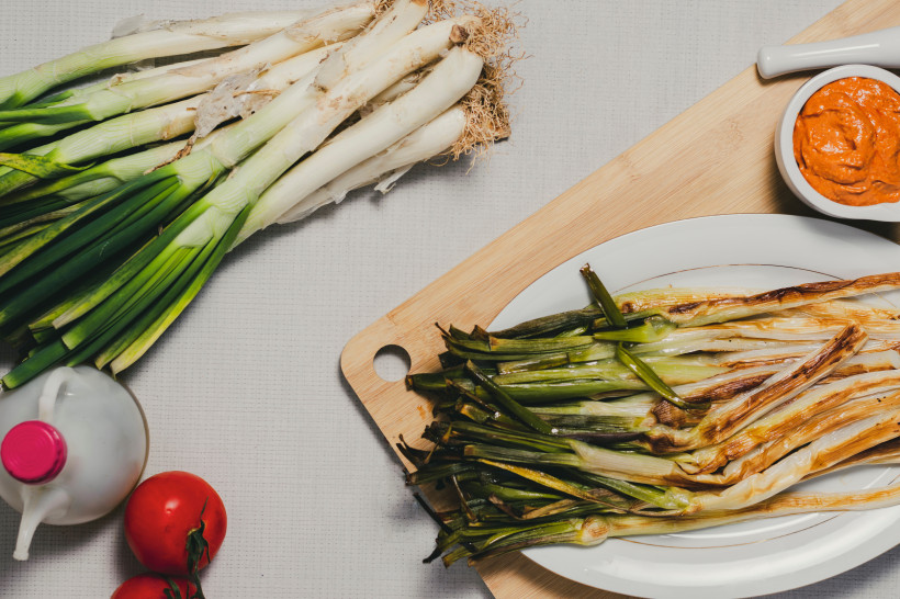 Frische und gegrillte Calçots auf Schneidebrett und Teller, daneben Schale mit roter Sauce