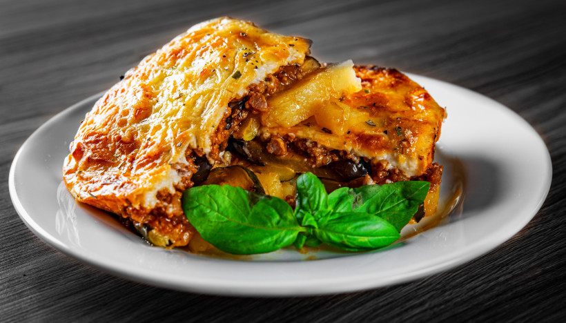 Stück Moussaka mit Kartoffel-, Auberginen- und Hackfleischschichten auf einem weißen Teller, mit Basilikum garniniert