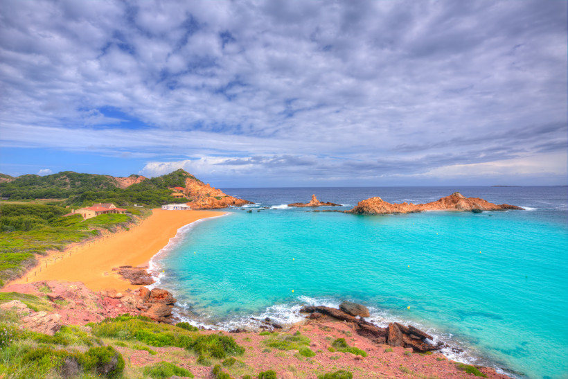 Cala Pregonda: Sandstrand, türkisfarbenes Wasser und Felsen Bucht Cala Pregonda mit goldenem Sandstrand, türkisfarbenem Meer und Felsen unter bewölktem Himmel