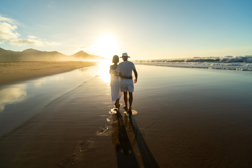 Fuerteventura Ein Paar in Weiß geht barfuß am Strand in den Sonnenuntergang. Sie halten sich im Arm, das Meer spiegelt das Licht der tief stehenden Sonne