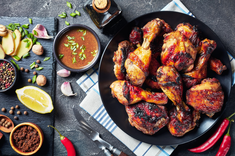 Jamaika Ein schwarzer Teller voller gegrillter Jerk Chicken Drumsticks, die außen knusprig und kräftig gewürzt sind. Daneben liegen auf einer Schieferplatte frische Zutaten wie Ingwer, Knoblauch, Zitronenspalten, Chili, Pfefferkörner, Muskatnuss und brauner Zucke
