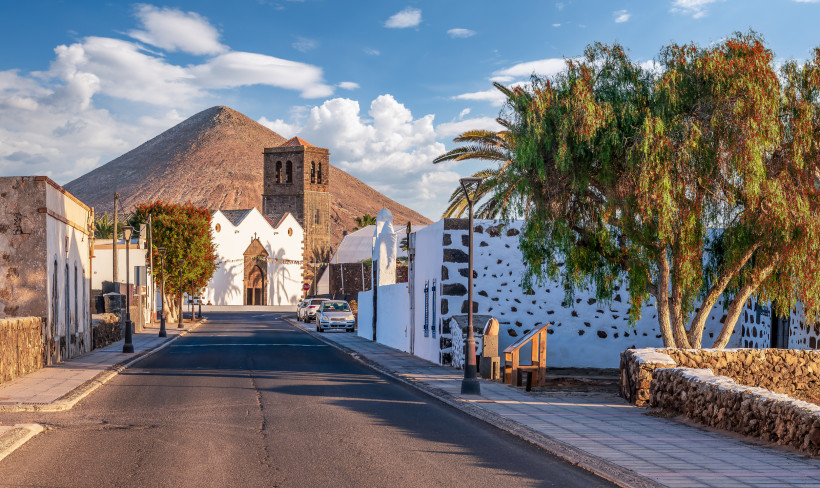 Fuerteventura Ruhige Straße mit weiß getünchten Häusern und Palmen, im Hintergrund eine historische Kirche mit Glockenturm und ein kegelförmiger Vulkan unter blauem Himmel mit Wolken.