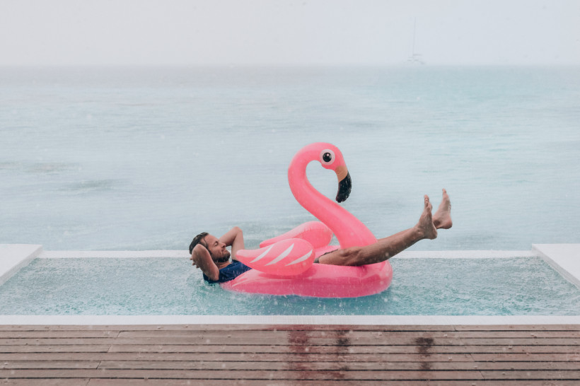Sommer in Mexiko Mann entspannt auf einem pinken Flamingo-Schwimmring in einem Infinity-Pool mit Blick auf das Meer, während leichter Regen fällt