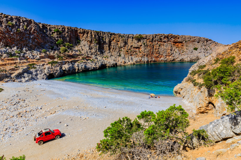 Bucht auf Kreta mit Kiesstrand, türkisblauem Wasser und Felsklippen, unten ein roter Geländewagen