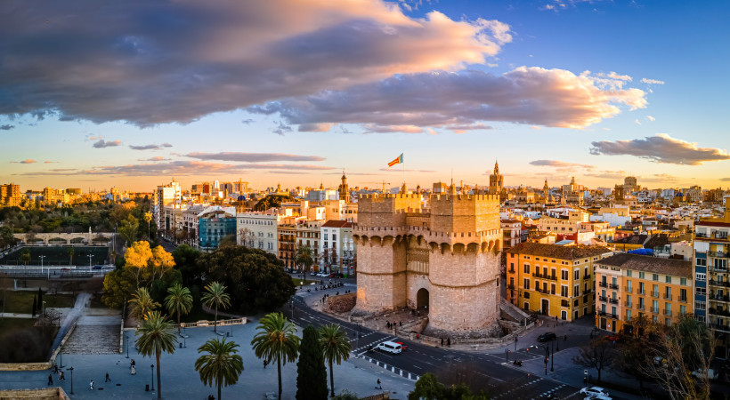 Panoramablick auf Valencia bei Sonnenuntergang. Im Vordergrund das imposante Stadttor Torres de Serranos mit wehender Flagge. Umgeben von Palmen, Altstadtgebäuden und dem ehemaligen Flussbett des Turia-Parks.