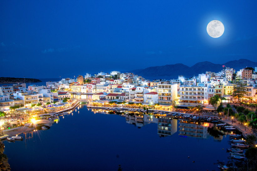 Abendansicht von Agios Nikolaos mit beleuchtetem Hafenbecken und Vollmond über der Küste