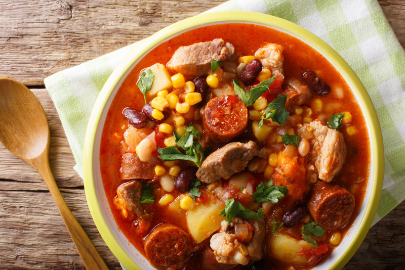 Nahaufnahme eines kapverdischen Eintopfs in einem tiefen Teller. Der Eintopf enthält Fleischstücke, Chorizo-Wurst, Mais, Bohnen, Kartoffeln und Tomaten in einer roten Brühe, garniert mit frischer Petersilie. Daneben liegt ein Holzlöffel auf einem karierte