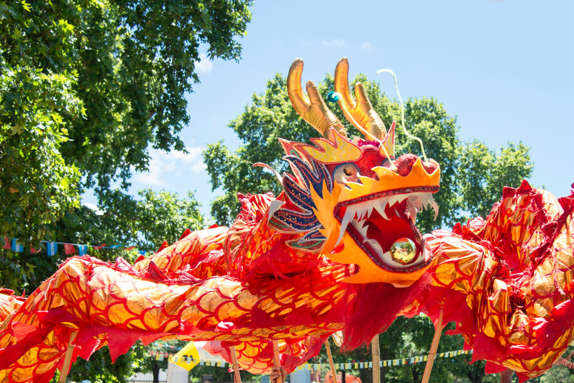 Thailand - Koh Samui Das Bild zeigt einen großen, farbenprächtigen chinesischen Drachentanz bei Tageslicht. Der Drache ist kunstvoll gestaltet in leuchtendem Rot, Gold und Orange, mit schuppigem Körper, wilden Augen, offenem Maul und goldener Kugel. Er wird von Stangen getrag