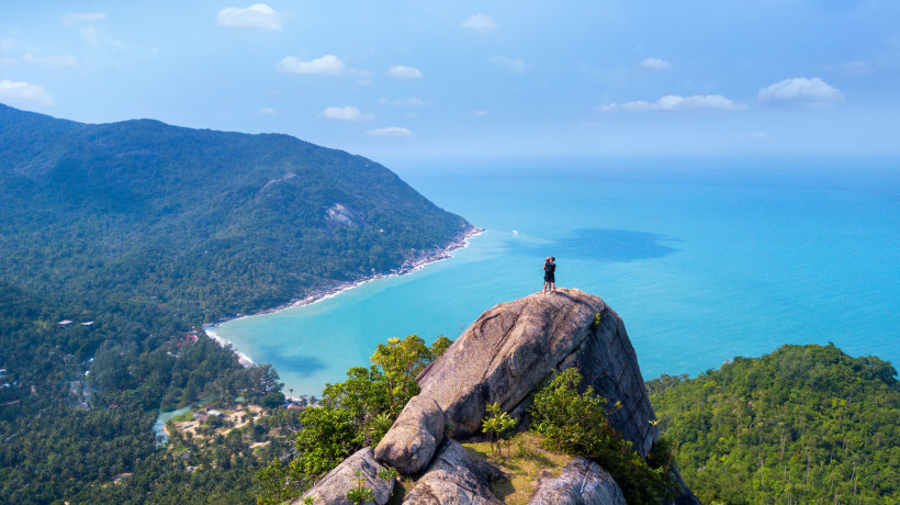 Person steht auf einem hohen Felsen mit weitem Blick über Meer und bewaldete Küste. ( Koh Phangan Island )