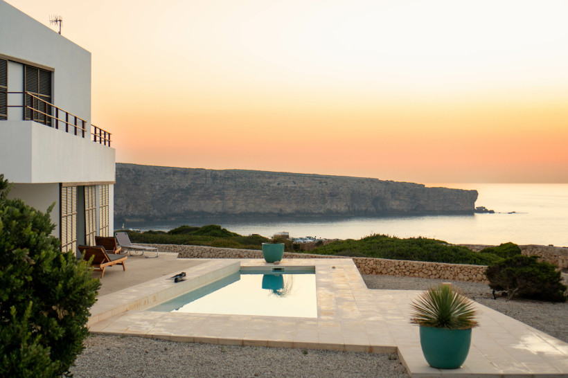 Moderne weiße Villa mit Poolterrasse auf Menorca bei Sonnenuntergang. Der Infinity-Pool spiegelt den Himmel in zarten Orange- und Gelbtönen wider. Im Hintergrund erhebt sich eine flache Steilküste über dem ruhigen Meer. Umgeben ist die Anlage von Steinmau