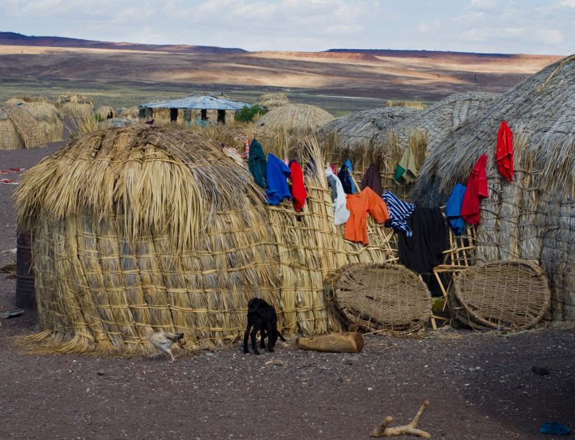 Kenia Traditionelle Rundhütten aus Schilf oder Gräsern mit gewölbten Dächern in einem ländlichen Gebiet. Über die Hütten ist bunte Kleidung zum Trocknen gehängt. Auf dem Boden laufen ein kleines schwarzes Zicklein und ein Huhn umher