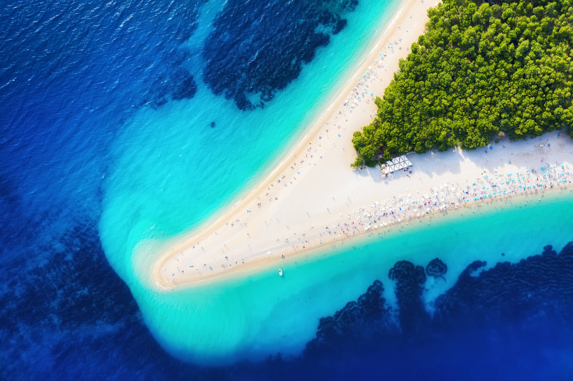 Draufsicht auf den berühmten Strand Zlatni Rat auf der kroatischen Insel Brač. Die schmale, sichelförmige Landzunge mit hellem Kiesstrand ragt ins türkisfarbene Meer, umgeben von dichtem Pinienwald und Badegästen mit bunten Sonnenschirmen.