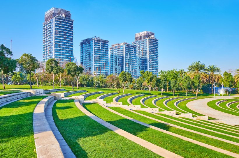Modern gestaltete Grünanlage mit runden Terrassen aus Rasen und Stein, die an ein Amphitheater erinnern. Im Hintergrund ragen mehrere moderne Hochhäuser in den Himmel. Palmen und Bäume säumen den Park, der Himmel ist klar und blau.