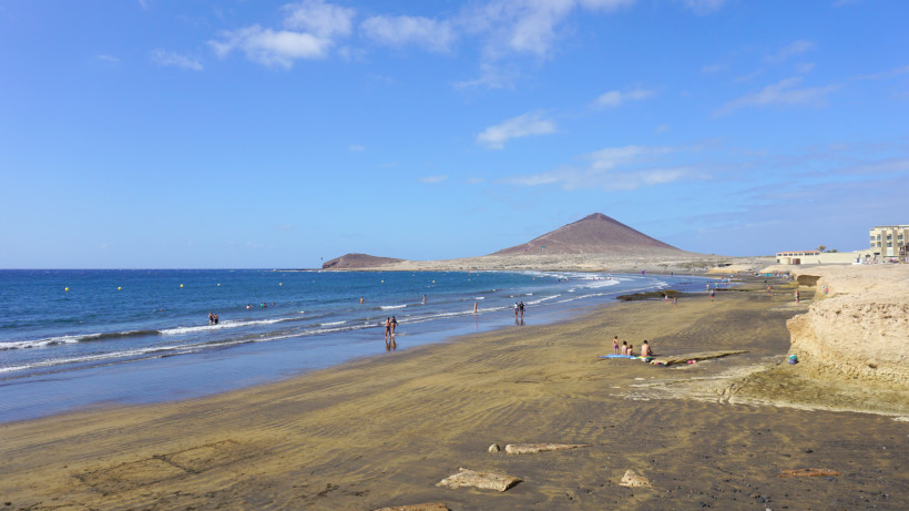 El Médano Strand mit Montaña Roja im Hintergrund Strand von El Médano auf Teneriffa mit flachem Wasser, Spaziergängern am Ufer und Blick auf die Montaña Roja unter blauem Himmel.