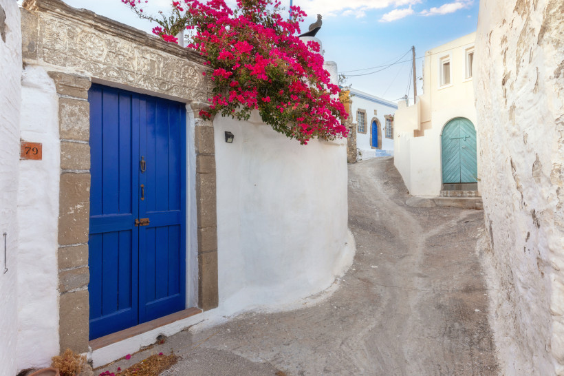 Schmale griechische Gasse mit weiß getünchten Häusern, blauer Holztür und pinker Bougainvillea in einem Dorf auf Rhodos