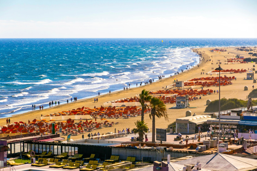Blick auf den Strand von Maspalomas mit Wellen, Spaziergängern und Reihen orangefarbener Sonnenschirme