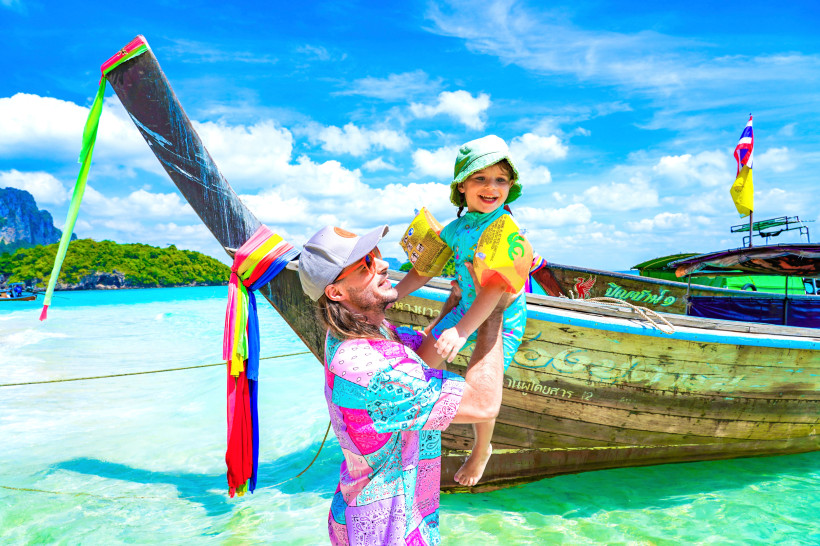 Thailand - Krabi Das Bild zeigt einen Vater, der sein kleines Kind lachend in die Luft hebt. Beide stehen im seichten, türkisfarbenen Wasser vor einem traditionellen thailändischen Langboot, das mit bunten Stoffbändern und Flaggen geschmückt ist. Der Vater trägt ein farbe