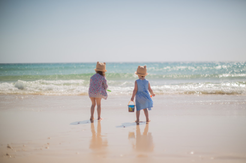 Zwei Kinder mit Sonnenhüten am Strand von Corralejo, mit Eimer und Spielzeug am Meeresufer