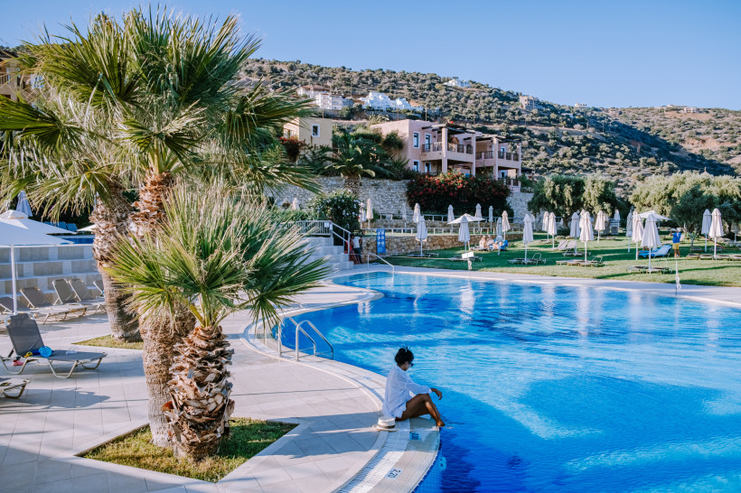 Hotelpool mit Liegen, Sonnenschirmen und Palmen vor Hügellandschaft; Person sitzt am Beckenrand