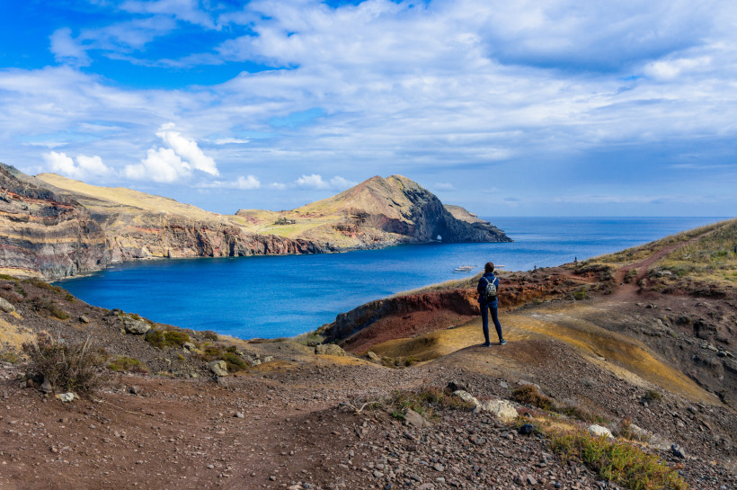 Madeira Person mit Rucksack blickt auf eine zerklüftete Küste mit türkisblauem Wasser auf Madeira. Der Wanderweg führt über farbige Felsen und Hänge, während sich im Hintergrund eine markante Landzunge ins Meer erstreckt. Der Himmel ist leicht bewölkt, aber freun