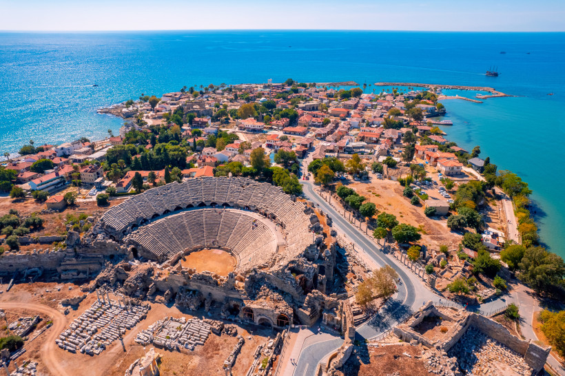 Das Bild zeigt eine Luftaufnahme von Side an der türkischen Riviera. Im Vordergrund liegt ein gut erhaltenes römisches Amphitheater, dahinter eine Altstadt mit roten Dächern auf einer schmalen Landzunge im Mittelmeer. Ruinen, enge Gassen und ein kleiner H