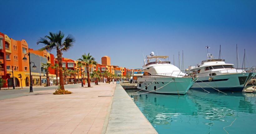 Das Bild zeigt die lebendige Marina von Hurghada am Roten Meer. Rechts im Bild liegen mehrere luxuriöse Yachten und Boote im türkisfarbenen Wasser vertäut. Links verläuft eine breite, gepflegte Promenade mit Palmen, Blumenbeeten und farbenfrohen Gebäuden 
