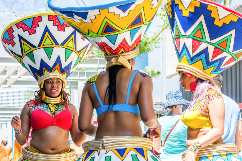 Südafrika Drei Frauen in auffälligen, bunten traditionellen Kostümen und riesigen, farbenfrohen Kopfbedeckungen tanzen bei einem Straßenfest. Sie tragen bunte BH-Oberteile und breite Schmuckreifen um Taille und Hals.