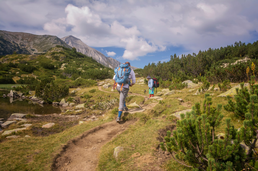 Zwei Wanderer mit großen Rucksäcken und Trekkingstöcken folgen einem schmalen Pfad durch die grüne Berglandschaft des Pirin-Gebirges in Bulgarien. Umgeben von Felsen, Sträuchern und einem kleinen Bergsee, ragen im Hintergrund schroffe Gipfel unter einem t