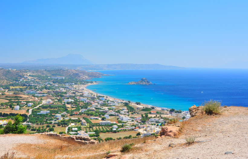 Kos Panoramablick auf eine griechische Küstenlandschaft mit klarem, tiefblauem Meer. Im Vordergrund liegt ein trockenes, leicht hügeliges Gelände mit Felsen und Sträuchern. Dahinter erstreckt sich ein Ort mit weißen Häusern entlang eines langen Sandstrands. D