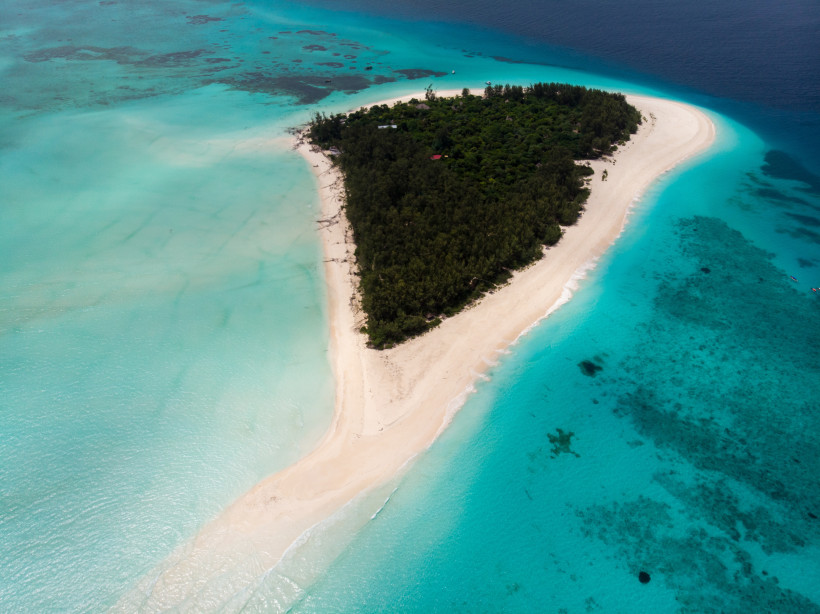 Sansibar - Mnemba Island  Luftaufnahme einer kleinen Insel vor Sansibar mit dichtem Grün und heller Sandbank, umgeben von türkisfarbenem Wasser.