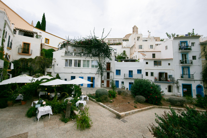 Häuserzeile in Dalt Vila mit weiß getünchten Fassaden, blauen Fensterläden und kleinem Platz mit Sonnenschirmen und Gartenbereich