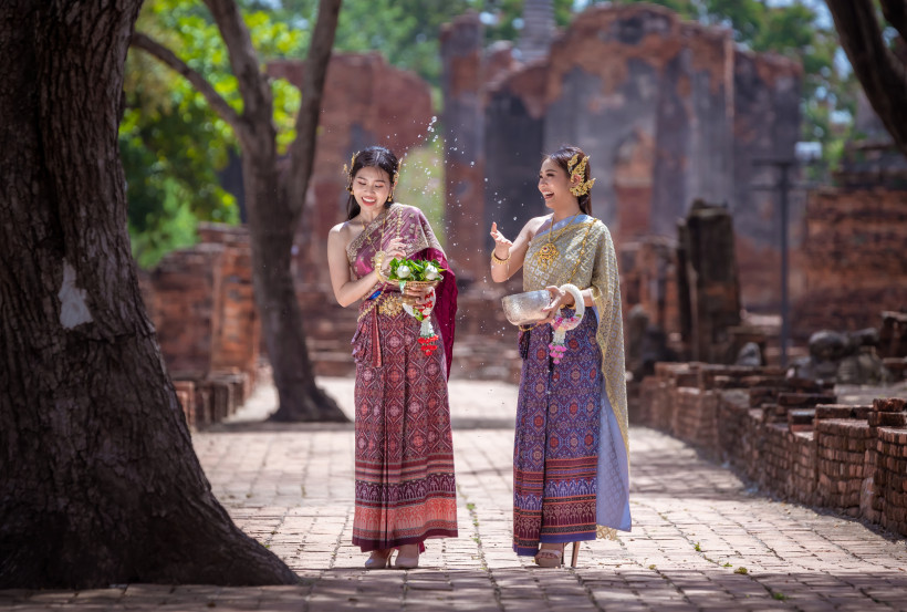 Thailand Zwei Frauen in traditioneller thailändischer Kleidung feiern lächelnd auf einem historischen Tempelgelände. Eine von ihnen spritzt Wasser aus einer Silberschale, die andere hält einen Blumen- und Blätterschmuck.