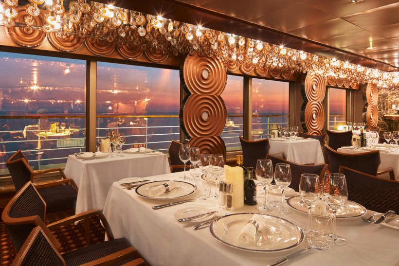 Innenansicht des Restaurants auf der Costa Favolosa mit Tischgedecken und Panoramafenstern zum Meer Restaurant an Bord der Costa Favolosa mit gedeckten Tischen, großen Fenstern und Blick auf das Meer bei Sonnenuntergang