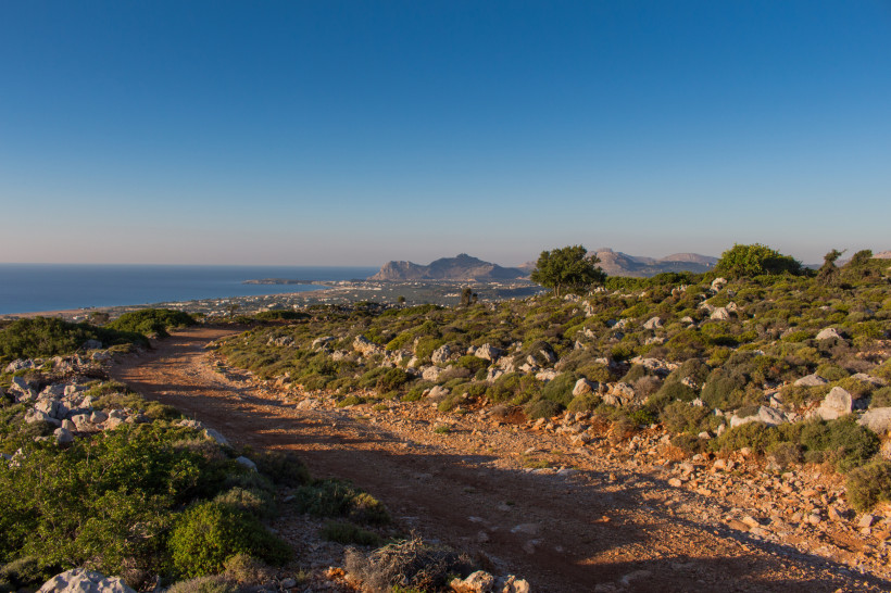 Kos - Psalidi Staubiger Wanderpfad mit Blick auf eine grüne, felsige Hügellandschaft und die weite Küste von Kos. Im Hintergrund liegen Bergketten, die von der untergehenden Sonne in warmes Licht getaucht werden.