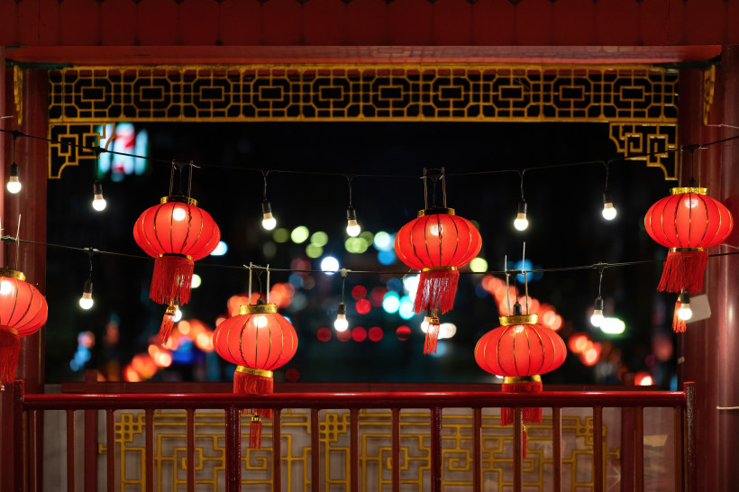 Thailand - Koh Samui Das Bild zeigt eine nächtliche Szene mit traditionellen roten chinesischen Laternen, die an schwarzen Kabeln zusammen mit kleinen Glühbirnen aufgehängt sind. Sie hängen vor einer verzierten Balustrade aus rotem Holz mit goldenen Mustern. Im Hintergrund le
