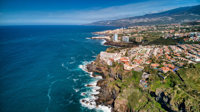 Dramatische Steilküste bei Puerto de la Cruz Atemberaubende Steilküste im Norden Teneriffas nahe Puerto de la Cruz, mit Wellen, die gegen dunkle Felsen schlagen. Rechts moderne Hotels und Häuser auf einer Klippe, dahinter grüne Terrassenlandschaft und Berge unter klarem Himmel.