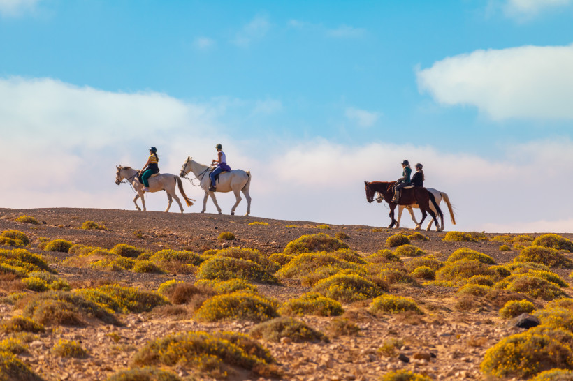 Fuerteventura Vier Menschen reiten auf Pferden durch eine trockene, hügelige Landschaft mit kleinen Sträuchern unter blauem Himmel mit hellen Wolken.