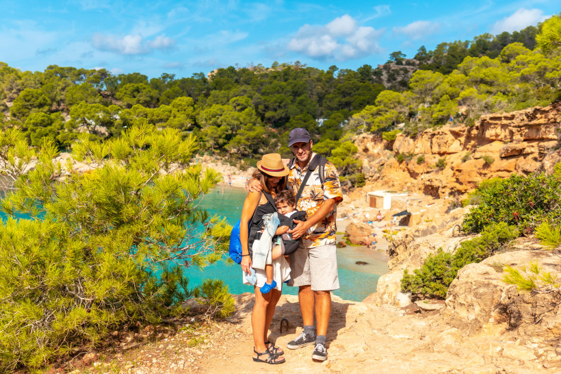 Ibiza Eine junge Familie steht an einem Aussichtspunkt über einer türkisblauen Bucht auf Ibiza. Die Eltern halten ein Kleinkind im Arm, alle lächeln in die Kamera. Im Hintergrund sind Felsen, grüne Pinien und das klare Wasser zu sehen. Die Szene vermittelt Urla